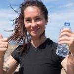 Happy woman giving thumbs up with water bottle on a sunny beach day in Portugal.