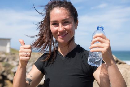 Happy woman giving thumbs up with water bottle on a sunny beach day in Portugal.